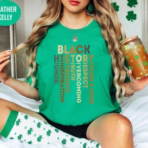 May include: A green t-shirt with the words "Black History" in brown and gold lettering. The words are arranged vertically in a column, with each word offset slightly from the one above it. The words "Ours & Mine", "Respect", "Overcoming", "Truth", "Inspiration", "Strength", "Honor" are also included in the design.