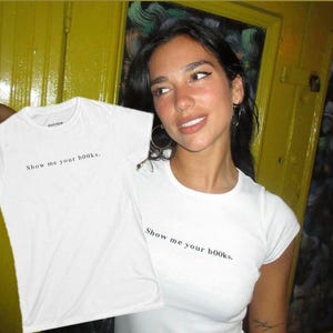Puede incluir: Camiseta blanca con el texto "Show me your books." La camiseta es de cuello redondo. La imagen muestra la camiseta levantada. El fondo es una pared amarilla.