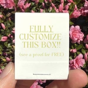 May include: A white rectangular matchbox with gold text that reads FULLY CUSTOMIZE THIS BOX!! (see a proof for FREE). The box features a single metal staple at the bottom center and is held against a background of pink azalea flowers.