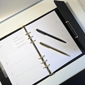 May include: An open black leather binder with a white planner insert. Two black pens and one gold pen are on the planner. The planner pages have lines for each day of the week and a habit tracker. A laptop and books are in the background.