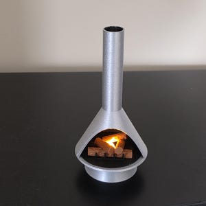 May include: A silver miniature fireplace with a cylindrical chimney and a conical firebox. Inside, a small pile of wooden logs glows with a warm, orange light. The fireplace is a decorative item, designed to create a cosy atmosphere.