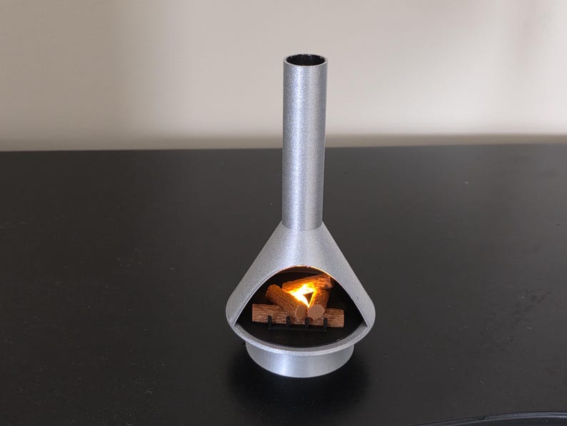 May include: A silver miniature fireplace with a cylindrical chimney and a conical firebox. Inside, a small pile of wooden logs glows with a warm, orange light. The fireplace is a decorative item, designed to create a cosy atmosphere.