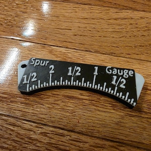May include: A black and white gauge tool with the text "Spur 2 1/2 1 Gauge 1/2". The tool has a curved shape and is marked with measurements in inches. It is designed for measuring and gauging.