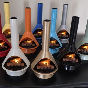 May include: A selection of miniature, brightly coloured fireplaces in a retro design. Each has a conical base, a tall chimney, and a simulated fire with logs. Colours include red, yellow, orange, blue, silver, black, and gold.