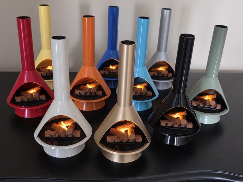 May include: A selection of miniature, brightly coloured fireplaces in a retro design. Each has a conical base, a tall chimney, and a simulated fire with logs. Colours include red, yellow, orange, blue, silver, black, and gold.