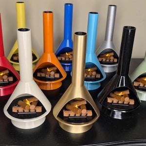 May include: A selection of brightly coloured, mid-century modern tabletop fireplaces. Each fireplace is a different colour, including red, yellow, orange, blue, silver, black, gold, and green. The fireplaces have a distinctive shape with a tall chimney and a firebox with faux logs.