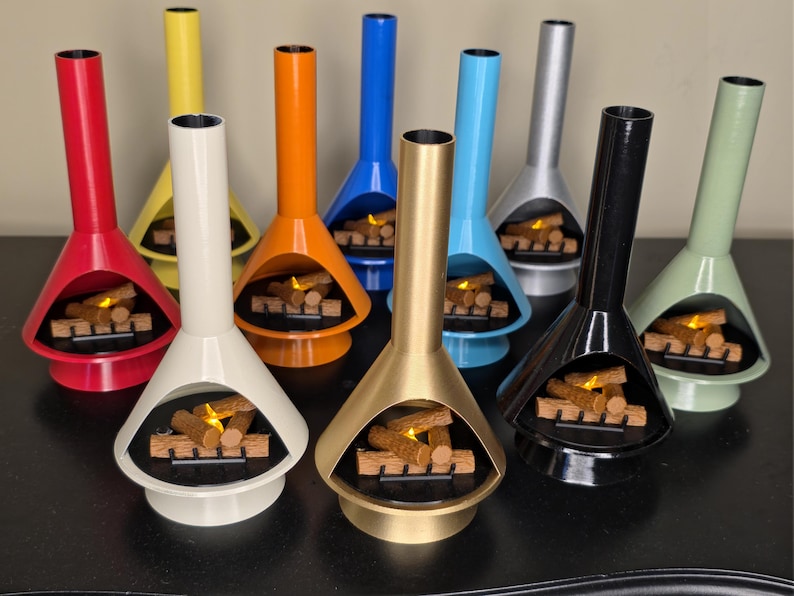 May include: A selection of brightly coloured, mid-century modern tabletop fireplaces. Each fireplace is a different colour, including red, yellow, orange, blue, silver, black, gold, and green. The fireplaces have a distinctive shape with a tall chimney and a firebox with faux logs.