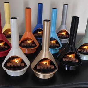 May include: A selection of colourful, retro-style tabletop fireplaces. Each fireplace has a conical body and a tall, cylindrical chimney. The fireplaces are in various colours, including red, yellow, orange, blue, silver, black, and green, with a glowing fire effect inside.
