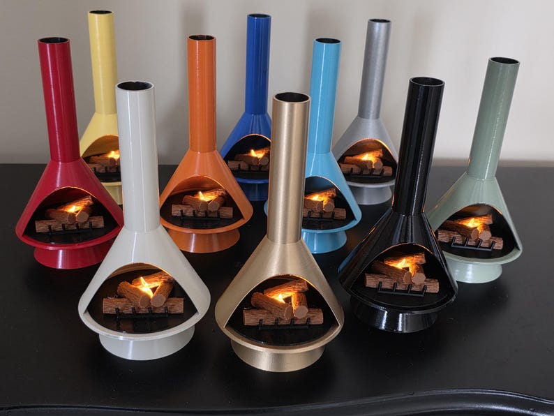 May include: A selection of colourful, retro-style tabletop fireplaces. Each fireplace has a conical body and a tall, cylindrical chimney. The fireplaces are in various colours, including red, yellow, orange, blue, silver, black, and green, with a glowing fire effect inside.