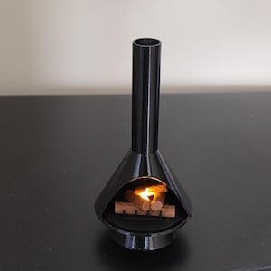 May include: A black, mid-century modern style miniature fireplace with a cylindrical chimney and a conical base. Inside the fireplace, there are small wooden logs with a flickering orange light, simulating a fire. The fireplace is on a black surface.