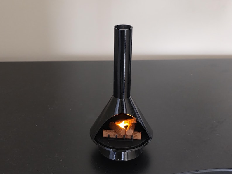 May include: A black, mid-century modern style miniature fireplace with a cylindrical chimney and a conical base. Inside the fireplace, there are small wooden logs with a flickering orange light, simulating a fire. The fireplace is on a black surface.