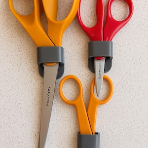 May include: Three pairs of Fiskars scissors in different sizes and colours. The scissors have orange and red handles with grey blade protectors. The blades are silver and the brand name "FISKARS" is printed on the blades.