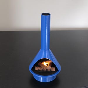 May include: A blue, mid-century modern fireplace with a cylindrical chimney and a flared base. Inside, a small stack of wooden logs with a flickering flame. The fireplace is made of metal. The height is approximately 20 cm.