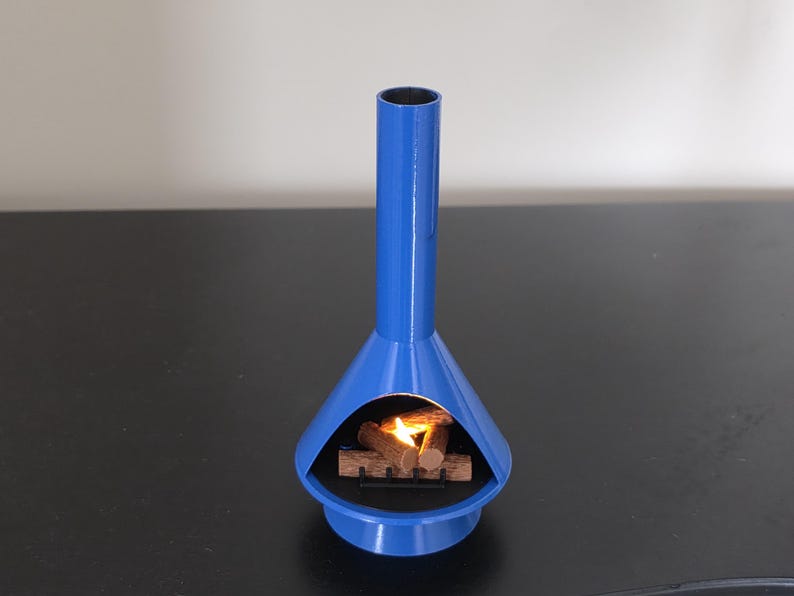 May include: A blue, mid-century modern fireplace with a cylindrical chimney and a flared base. Inside, a small stack of wooden logs with a flickering flame. The fireplace is made of metal. The height is approximately 20 cm.