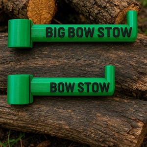 May include: Two bright green bow storage devices with the text "BIG BOW STOW" and "BOW STOW" in black. The devices are resting on a piece of wood. The background includes more wood and some greenery.