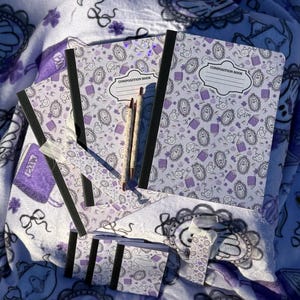 May include: A collection of purple and white stationery items, including composition notebooks, pencils, and a bookmark, arranged on a matching patterned blanket. The notebooks have black spines and the text "COMPOSITION BOOK". The blanket and stationery feature a repeating design of ghosts and other spooky elements.