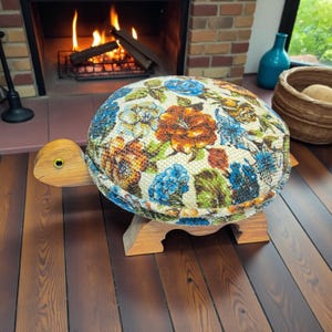 May include: A turtle-shaped footstool featuring a wooden head and four wooden legs. The shell is upholstered in a textured fabric with a colorful floral pattern in shades of orange, blue, and green on a cream background, with a yellow eye.