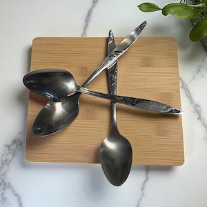 May include: Four silver spoons rest on a light brown bamboo cutting board. The spoons have a classic design, with a rounded bowl and a handle with a floral pattern. The board sits on a white marble surface, with a green plant in the background.