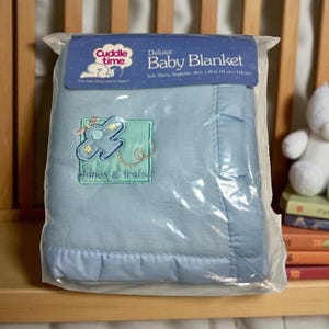 May include: Deluxe baby blanket in a plastic package. The light blue blanket has a stitched border and an embroidered design of a plane and stars on a green square. The blanket measures 36 x 45 inches.