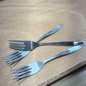May include: Three stainless steel forks with a floral rose design on the handles. The forks are arranged on a light brown wooden surface. The tines of the forks are facing different directions.