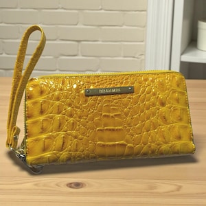 May include: A yellow, textured leather wallet with a wrist strap. The wallet has a zipper closure and a small metal plate with the brand name "BRAHMIN". The leather has a crocodile-like pattern.