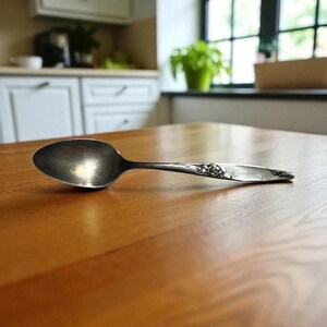 May include: A silver spoon with a floral design on the handle rests on a wooden table. The spoon has a rounded bowl and a detailed, decorative handle. The spoon is a kitchen utensil.