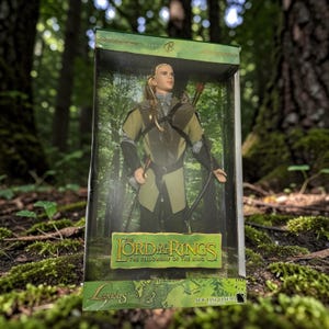 May include: A Legolas action figure from The Lord of the Rings: The Fellowship of the Ring, in its original packaging. The figure is dressed in a green and black outfit and holds a bow and arrow. The packaging features the movie's logo and a map.