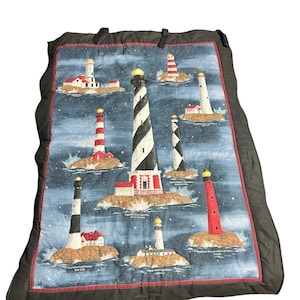 May include: A quilted blanket featuring illustrations of various lighthouses against a blue background. The lighthouses are depicted in different colors and designs, set on small islands with water splashing around them. The blanket has a dark border.