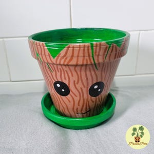 May include: A terracotta flower pot painted with a brown and green pattern to resemble the character Groot from the Guardians of the Galaxy movie franchise. The pot has a green rim and a green saucer.
