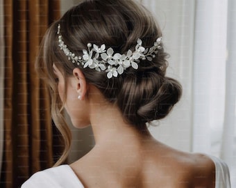 May include: A bridal hair accessory featuring a delicate floral design. The headpiece is adorned with white flowers, pearls, and crystals, set on a gold-toned wire. The accessory is worn on the back of the head, above a low bun.
