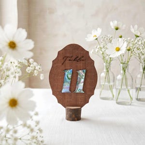 May include: A dark wood table number sign with a scalloped edge, featuring the text "Table" in black script above the number "11" inlaid with iridescent abalone shell. The sign is mounted on a small, cylindrical natural wood base.