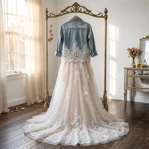 Pode incluir: Uma jaqueta jeans azul claro com detalhes em renda branca e o texto "Mrs. Smith" é exibida sobre um vestido de noiva branco e blush. O vestido é longo com uma sobreposição de tule e aplicações de renda floral. O conjunto está pendurado em um cabide de metal dourado.