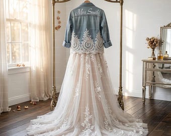 May include: A light blue denim jacket with white lace detailing and the text "Mrs. Smith" is displayed over a white and blush wedding dress. The dress is long with a tulle overlay and floral lace appliques. The ensemble hangs on a gold metal clothing rack.