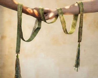 May include: A long, olive green fabric ribbon featuring a decorative gold-toned border pattern and finished with matching green and gold tassels at each end. The ribbon is draped in a series of loops, showcasing its texture and length.