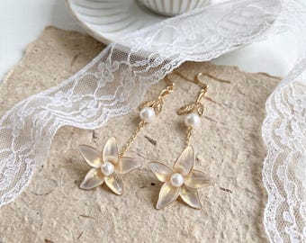 May include: A pair of gold-toned flower earrings with pearl accents. Each earring features a five-petal flower with a pearl center, suspended from a gold chain and a pearl. The earrings are set against a neutral background with lace.