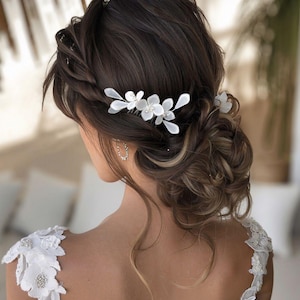 May include: A woman's updo hairstyle with a braided crown and a low bun. The hair is dark brown with blonde highlights. A white floral hair comb is placed in the updo. The woman is wearing a white dress with floral lace detailing.