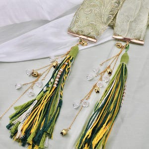May include: Two decorative tassels with green, yellow, and white threads. Each tassel has a green top, gold accents, and dangling chains with small white flower and bell charms. The tassels are attached to a light green ribbon.