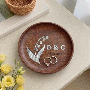 Könnte beinhalten: Ein runder Holztablett mit floralem Design, den Initialen "D & C" und dem Datum "2026.01.01" eingraviert. Zwei silberne Ringe liegen auf dem Tablett. Gelbe Rosen und ein geflochtener Korb sind im Hintergrund.