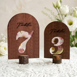 Könnte beinhalten: Zwei Tischkarten aus Holz. Jedes Schild hat eine dunkelbraune, gewölbte Oberseite mit dem Wort „Table“ in Schreibschrift. Die Zahlen 7 und 3 sind ausgeschnitten und zeigen ein Blumenmuster. Die Schilder sind auf kleinen Holzsockeln montiert.