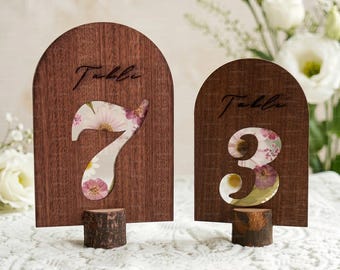 Könnte beinhalten: Zwei Tischkarten aus Holz. Jedes Schild hat eine dunkelbraune, gewölbte Oberseite mit dem Wort „Table“ in Schreibschrift. Die Zahlen 7 und 3 sind ausgeschnitten und zeigen ein Blumenmuster. Die Schilder sind auf kleinen Holzsockeln montiert.