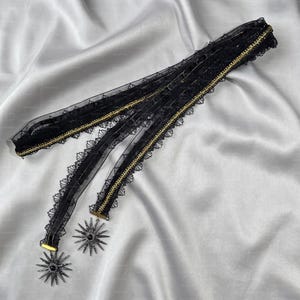 May include: A black lace choker featuring a sheer mesh ribbon with scalloped edges and a thin gold metallic trim. Two dangling starburst-shaped pendants with central black stones are attached to the ends of the ribbon, creating a layered effect.