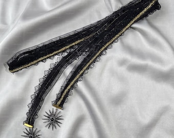 May include: A black lace choker featuring a sheer mesh ribbon with scalloped edges and a thin gold metallic trim. Two dangling starburst-shaped pendants with central black stones are attached to the ends of the ribbon, creating a layered effect.