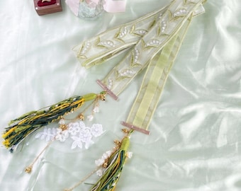 May include: A sheer light green ribbon featuring white embroidered lily of the valley flowers, finished with long tassels in shades of green, yellow, and white, accented with small gold-toned metal charms and white floral lace appliqués.