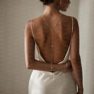 May include: A delicate gold-toned back necklace featuring a thin chain and small white pearls, worn with a sleeveless cream-colored satin slip dress that has a low-cut back design, thin shoulder straps, and a smooth, flowing fabric drape.