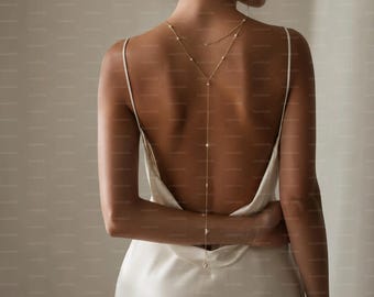 May include: A delicate gold-toned back necklace featuring a thin chain and small white pearls, worn with a sleeveless cream-colored satin slip dress that has a low-cut back design, thin shoulder straps, and a smooth, flowing fabric drape.