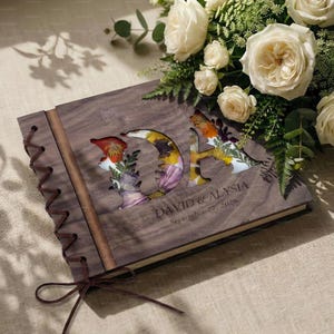 K&ouml;nnte beinhalten: Ein h&ouml;lzernes Hochzeitsg&auml;stebuch mit den ausgeschnittenen Initialen "DA", gef&uuml;llt mit bunten Trockenblumen. Das Buch ist mit einer Lederschnur gebunden und tr&auml;gt die Namen "DAVID & ALYSIA" und das Datum "27. September 2026".