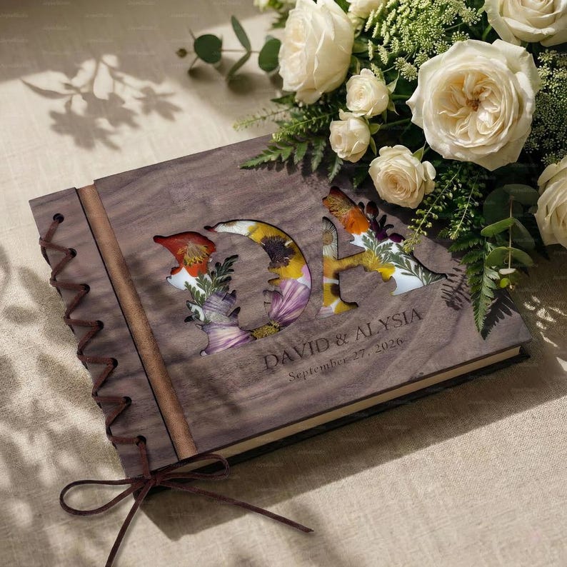 K&ouml;nnte beinhalten: Ein h&ouml;lzernes Hochzeitsg&auml;stebuch mit den ausgeschnittenen Initialen "DA", gef&uuml;llt mit bunten Trockenblumen. Das Buch ist mit einer Lederschnur gebunden und tr&auml;gt die Namen "DAVID & ALYSIA" und das Datum "27. September 2026".