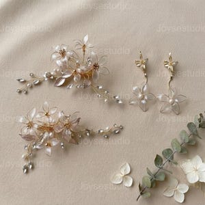 May include: A set of bridal hair accessories and earrings. The image shows two floral hair clips and a pair of dangle earrings. The hair clips and earrings feature delicate, translucent flowers, crystals, and pearls, all set on gold-toned wire.