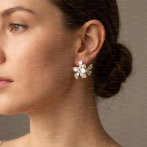 May include: Close-up of a woman wearing white floral earrings. Each earring features a cluster of delicate, petal-like shapes with a pearl-like centre. The earrings are a light, neutral colour, and the design is reminiscent of small flowers.