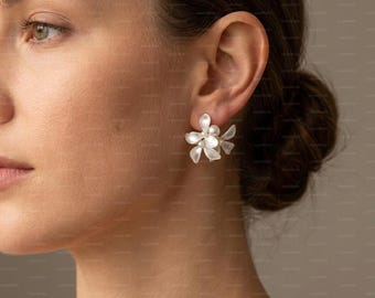 May include: Close-up of a woman wearing white floral earrings. Each earring features a cluster of delicate, petal-like shapes with a pearl-like center. The earrings are a light, neutral color, and the design is reminiscent of small flowers.
